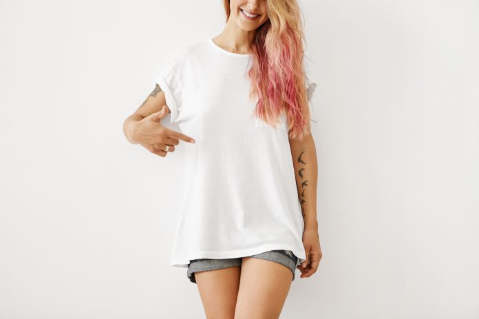 Clothing, design, style, fashion and advertising. Portrait of unrecognizable slim hipster girl with pink hair highlights and tattooed arms smiling, pointing finger at copy space on white black t shirt